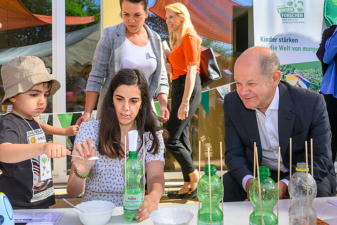 Ein Kind füllt Zitronensäue in eine Wasserflasche, eine Erzieherin hilft ihm dabei. Bundeskanzler Olaf Scholz beobachtet, was passiert.